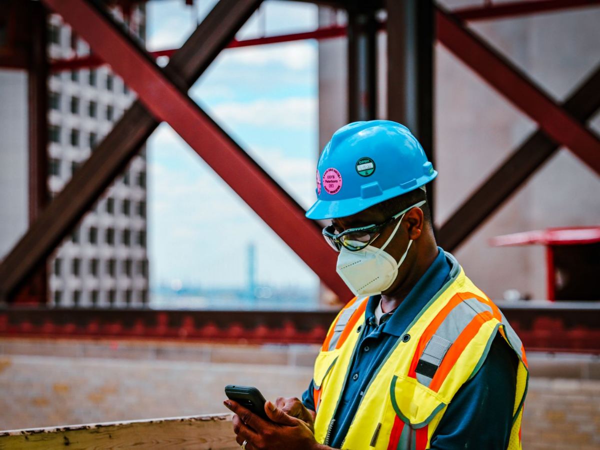 Worker in a hard hat, safety glasses, and hi-vis vest checking a phone on a steel-frame jobsite